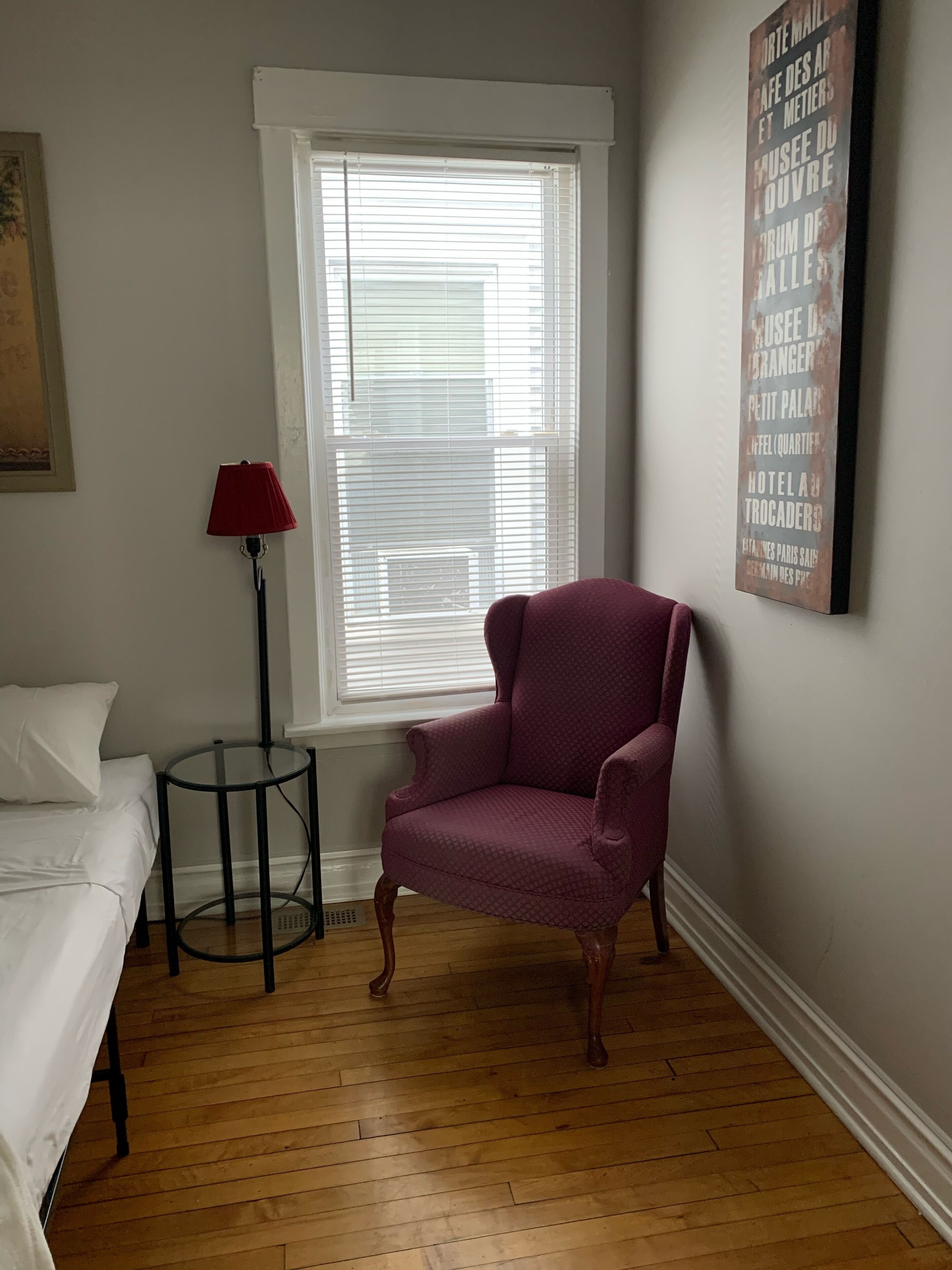A room with a wooden floor, a white bed, a red lamp, a purple armchair, and a window with blinds.