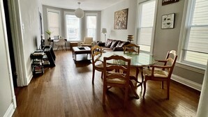 Un salón espacioso con pisos de madera, un comedor con una mesa redonda y sillas, y ventanas amplias con persianas.