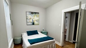 Un dormitorio con cama, mesita de noche, un cuadro y una puerta que da a otra habitación.