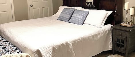 A neatly made bed with a white comforter and two pillows, a wooden headboard, a small bedside table with two candles, and a patterned rug on the floor.