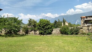 A grassy field with stone walls and trees, a stone building with a bell tower, and mountains in the background. - Holidayapartments in beautiful house, private garden, free Wi-Fi (Tignale)