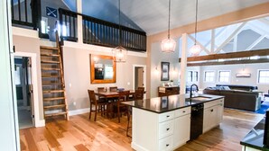 A spacious kitchen with wooden flooring, a central island, and a dining area with chairs. There is a staircase leading to an upper level.