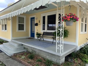 Una casa amarilla con puerta azul, número 209, un porche con escalones, un banco y una maceta colgante.