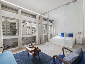 A modern bedroom with a large bed, a desk with a chair, a small table, and a view of the city through large windows. - Building in the Funchal centre, Drapes Design (Funchal)