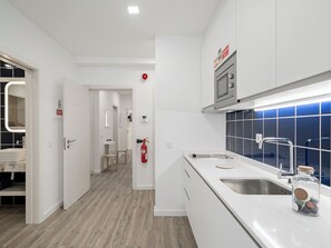 A modern kitchen with white cabinets, a stainless steel sink, and a microwave. There is a fire extinguisher mounted on the wall and a small table with chairs in the background. - Building in the Funchal centre, Drapes Design (Funchal)