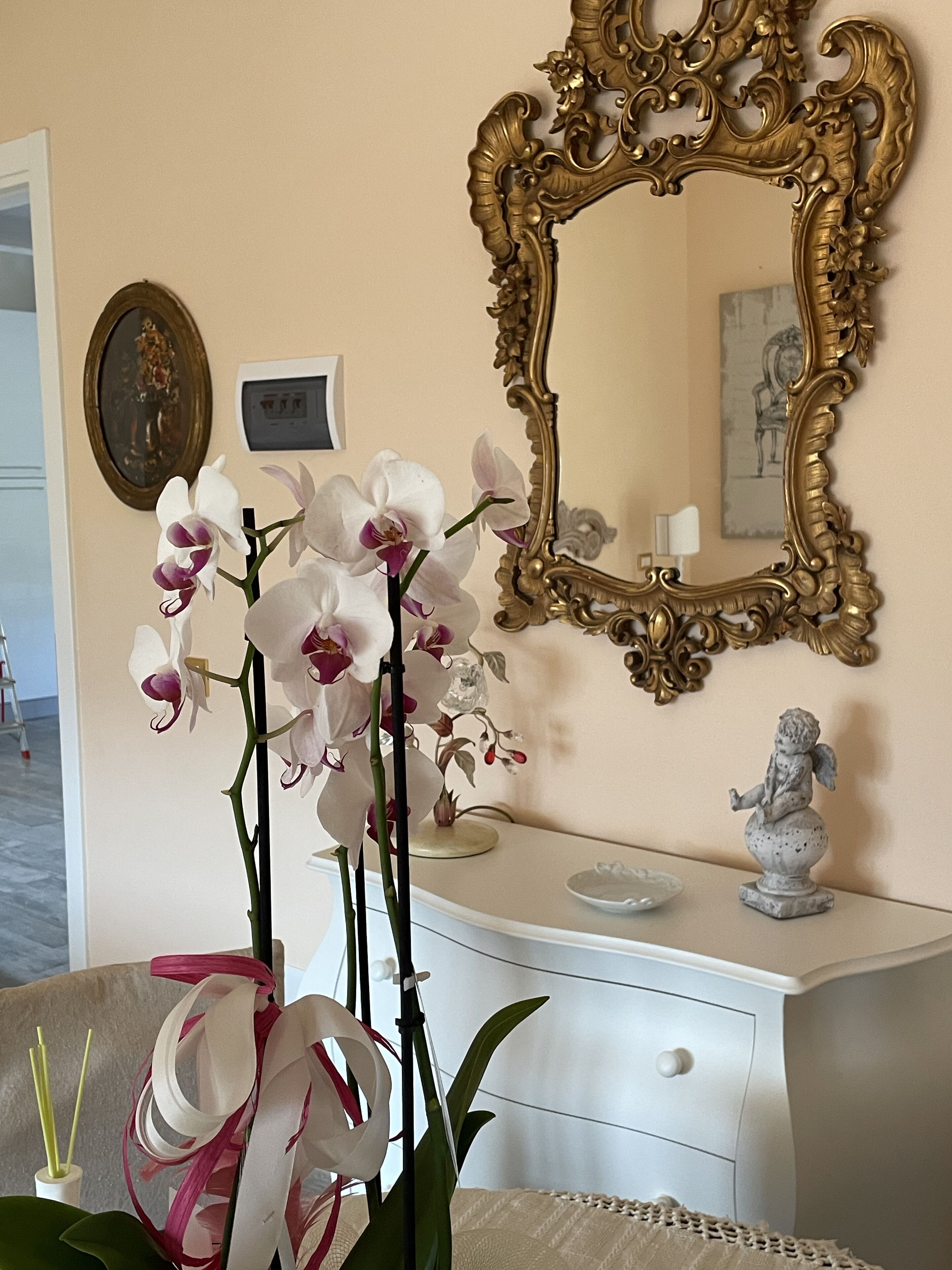 Una habitación con un tocador blanco, un espejo dorado ornamentado y orquídeas blancas.