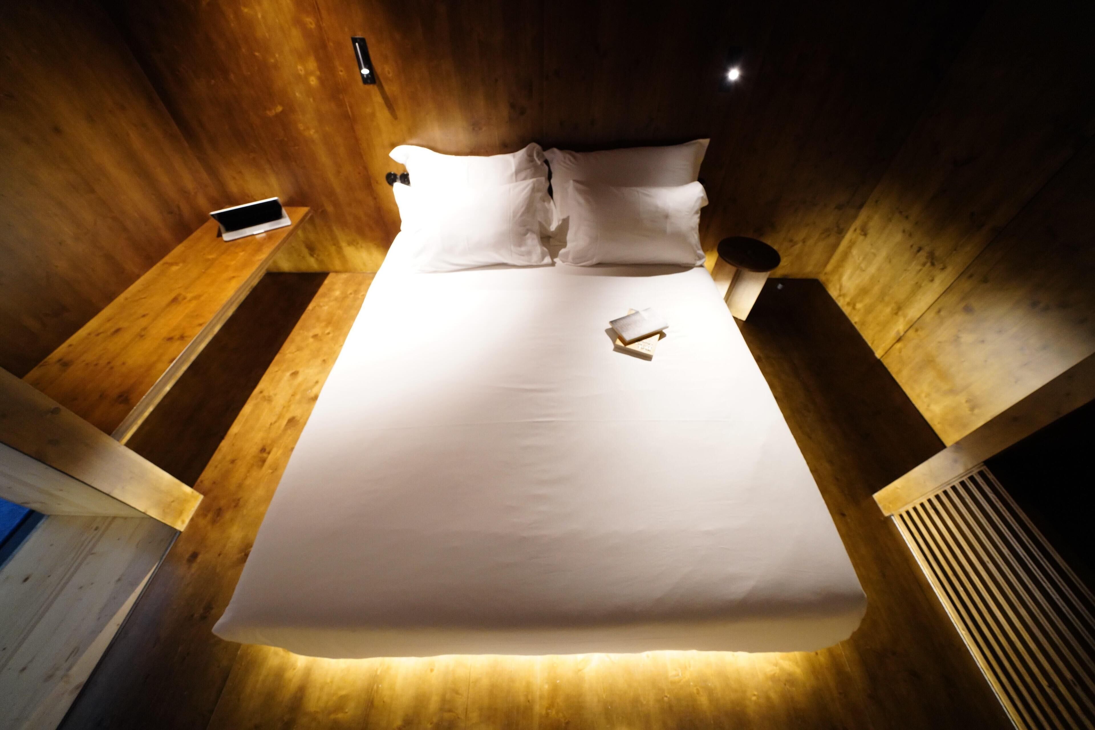 A neatly made bed with white linens and pillows, a bedside table with a smartphone, and a wall-mounted light fixture.