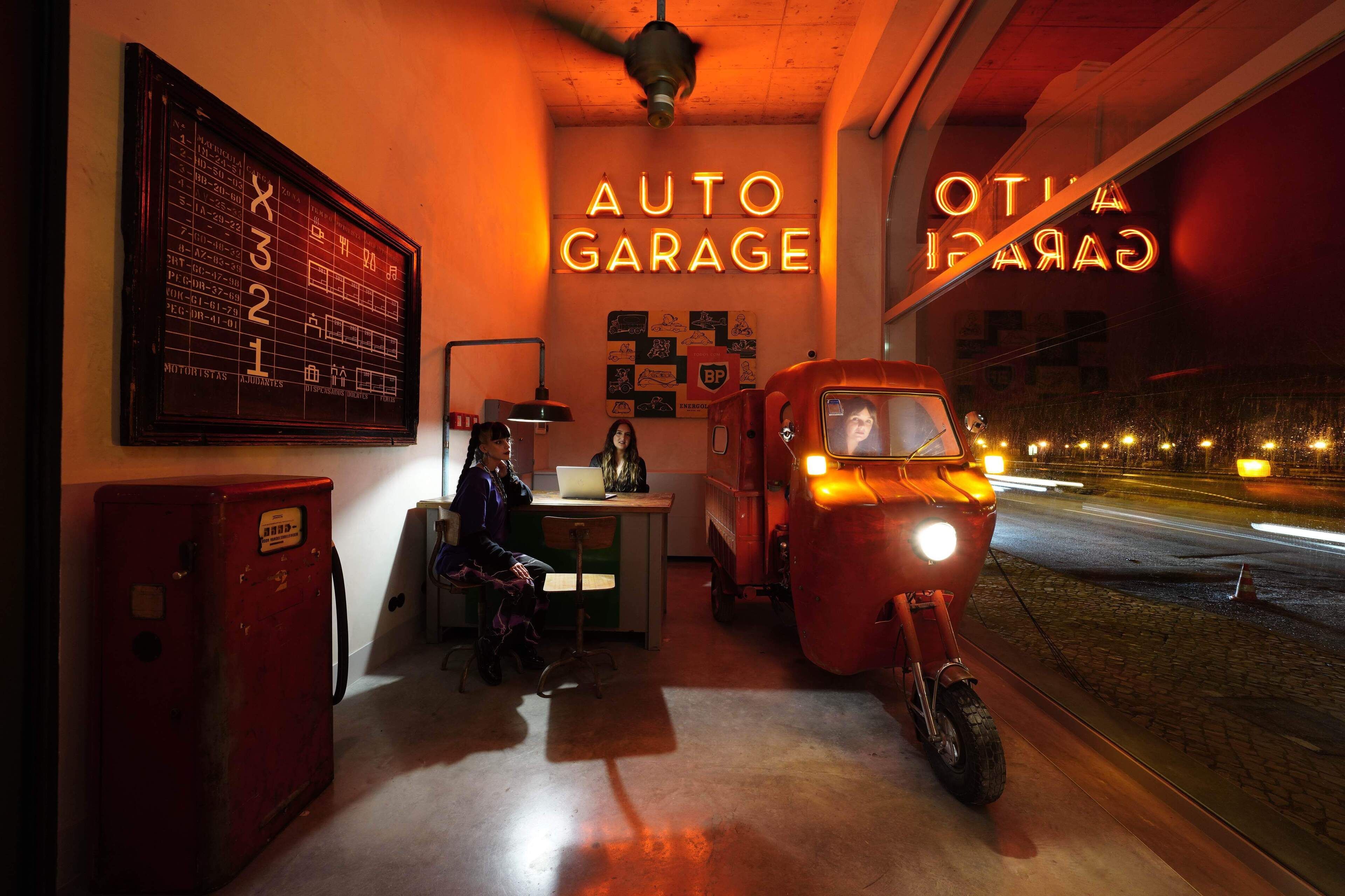 A vintage auto garage interior with a neon sign, a vintage car, and a motorcycle.