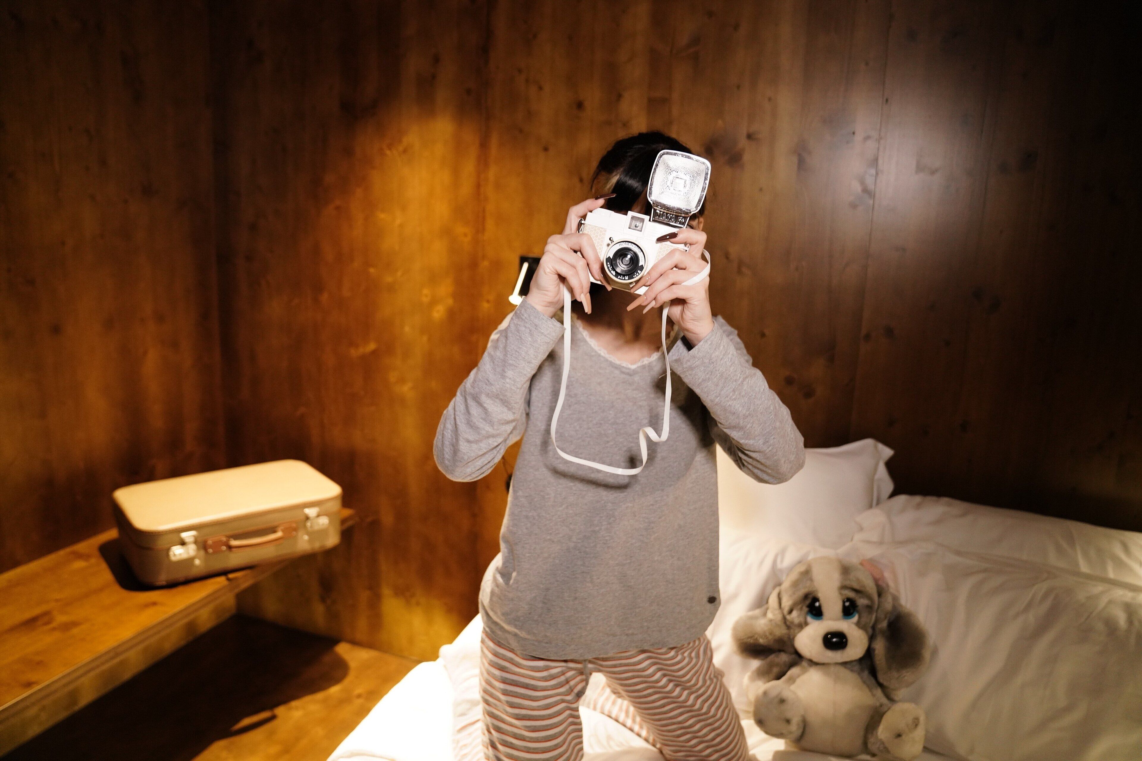 Une personne prend une photo avec un appareil photo ancien dans une pièce aux murs en bois, avec un lit recouvert de draps blancs et un petit chien sur le lit.