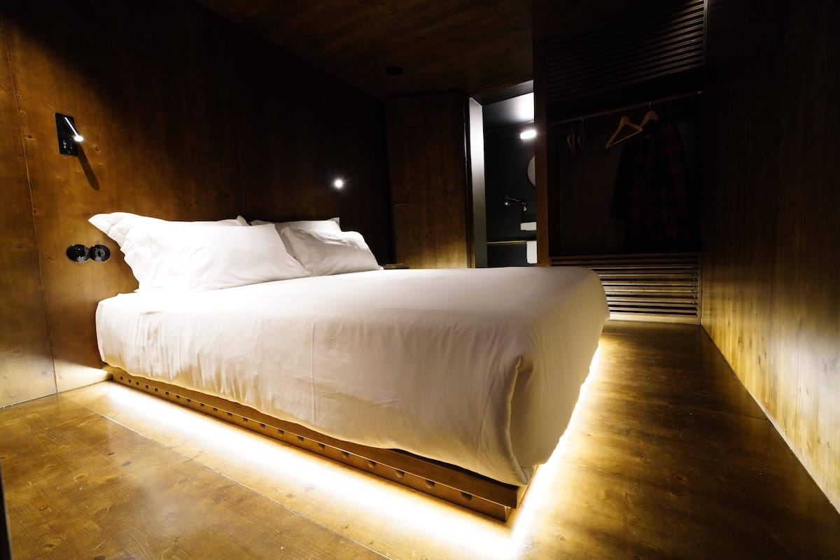 A modern bedroom with a wooden floor and walls, a bed with white bedding, a wall-mounted light, and a closet with a hanger.