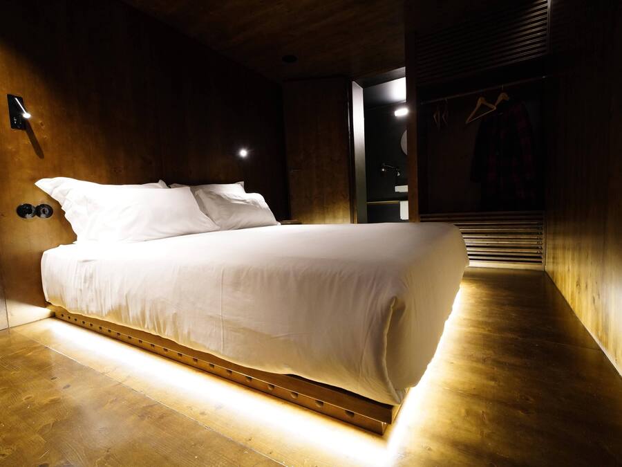 A modern bedroom with a wooden floor and walls, a bed with white bedding, a wall-mounted light, and a closet with a hanger.