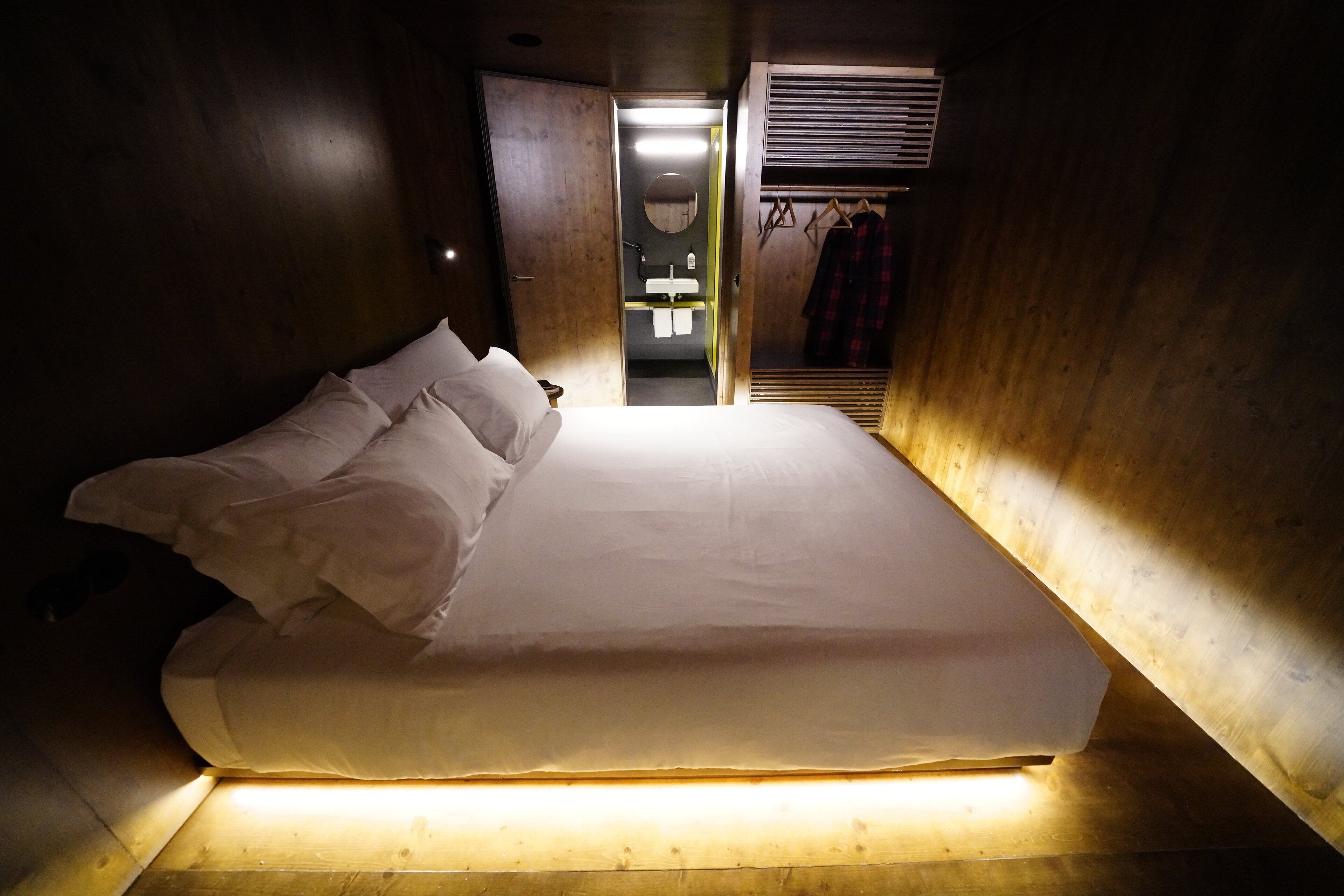 Een enkel bed met witte lakens in een kamer met houten wanden, met een kleine badkamer zichtbaar door een open deur.