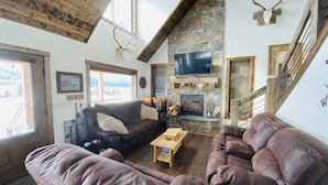 A cozy cabin interior with a stone fireplace, leather sofas, a wooden coffee table, and a TV mounted on the wall. - MODERN RUSTIC CABIN WITH WATER ACCESS AND BOAT DOCK [SLEEPS 17] (West Yellowstone)
