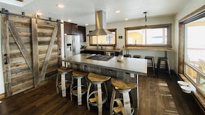 A kitchen with a central island, wooden bar stools, a large window, and a rustic wooden door. - MODERN RUSTIC CABIN WITH WATER ACCESS AND BOAT DOCK [SLEEPS 17] (West Yellowstone)