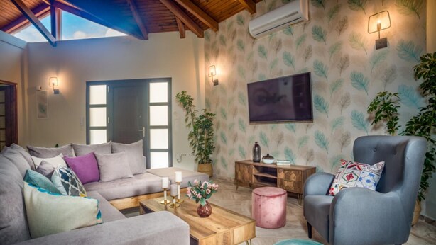 Un salon avec un canapé gris, un fauteuil bleu, une table basse en bois et un pouf rose. Un téléviseur est fixé au mur et une plante en pot est posée sur le sol.