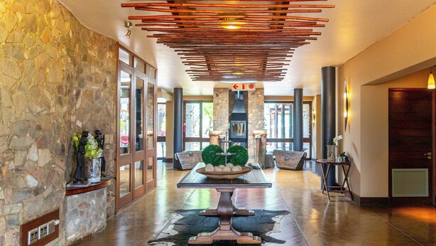 A spacious lobby with a stone wall, a wooden reception desk, a central fireplace, and a ceiling with wooden slats.