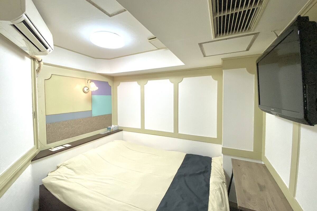 A hotel room with a bed, a flat-screen TV, an air conditioning unit, and a window with blinds.