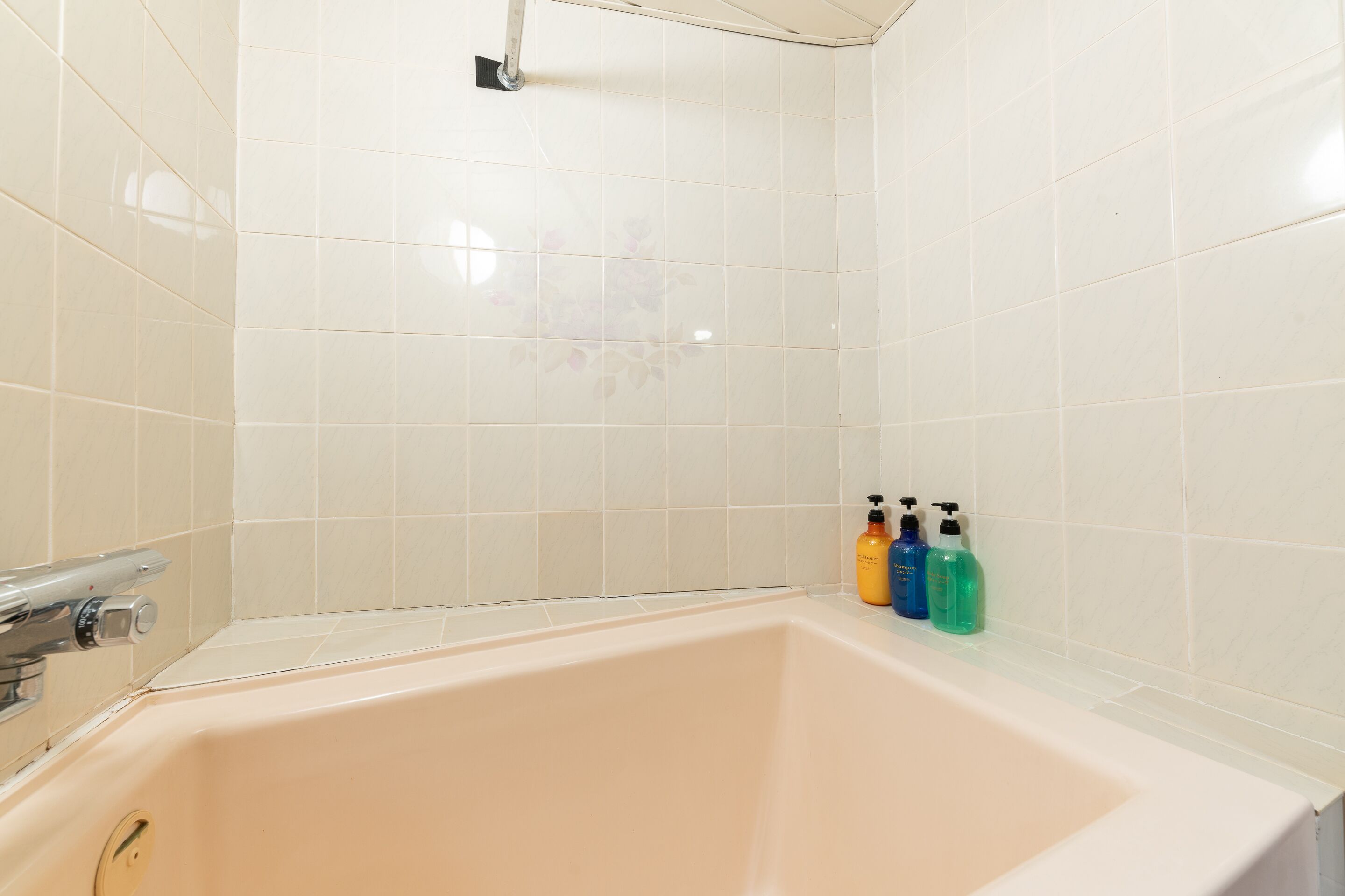 A bathroom with a bathtub, white tiles, and three bottles of toiletries on the edge.