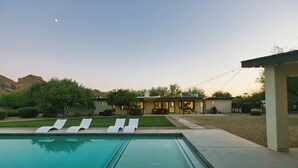 A swimming pool with lounge chairs, a patio area, and a house with string lights.