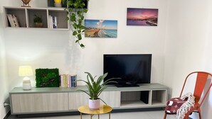 Una sala de estar moderna con una televisión de pantalla plana, una estantería, una mesita con una planta, un sillón rojo y una cama con colcha azul.