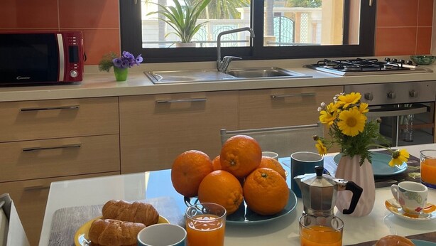 Una cocina moderna con un ventanal grande, un microondas, un fregadero y una estufa. Hay naranjas y pasteles en la encimera, y una mesa de centro con un mantel individual.