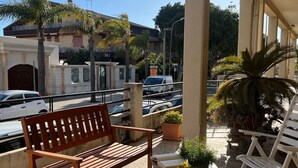 Un espacio de estar al aire libre cubierto con un banco de madera y una pequeña mesa, rodeado de plantas en macetas y vistas a una calle con coches y edificios.