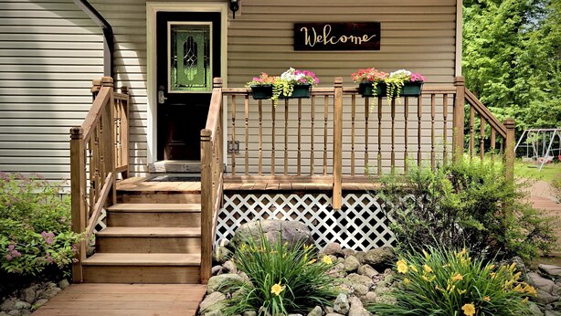 Een houten trap leidt naar een huis met een 'Welcome'-bord, een voordeur en bloembakken op de veranda.