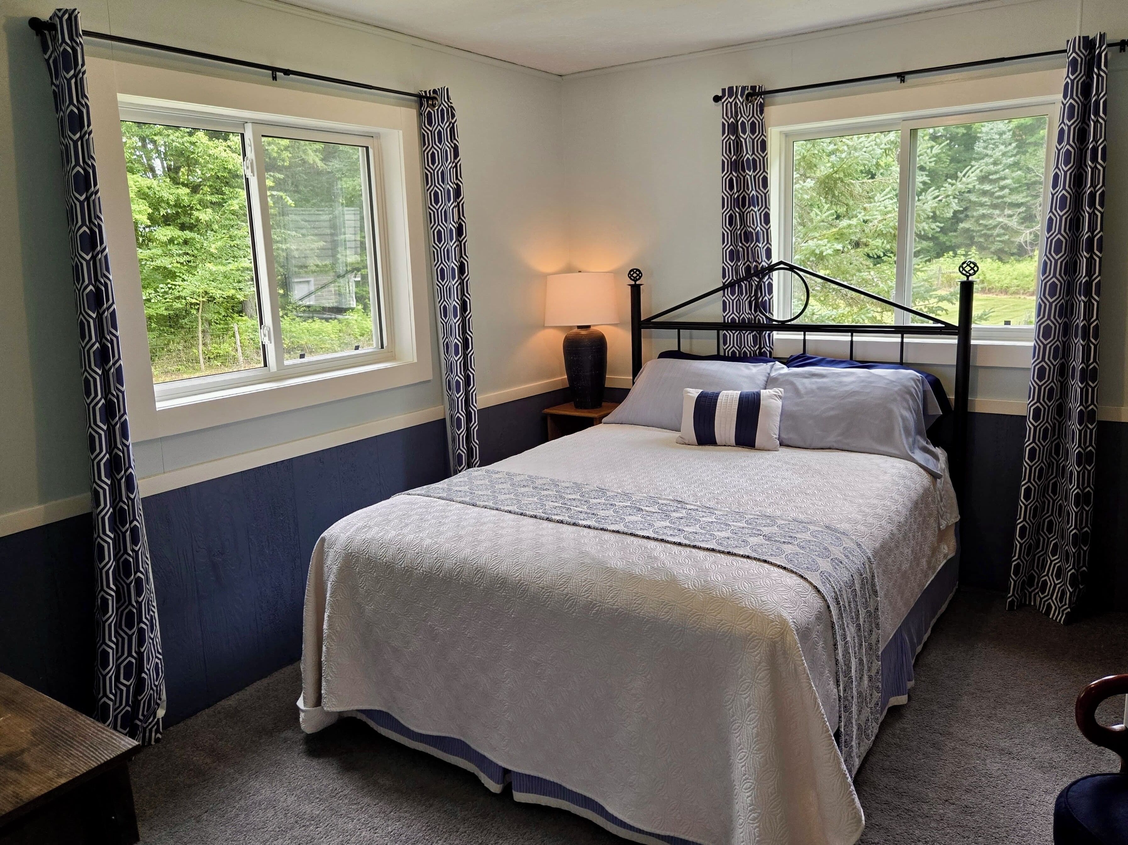 A bedroom with a bed, two windows, a nightstand, and a lamp.