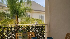 A table with a floral tablecloth, a carton of Solevita juice, and several blue cups on a balcony with a view of palm trees and a building. - Vacation home "Fortuna " (Balestrate)