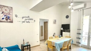 A living room with a blue sofa, a dining area with a table and chairs, a wall with a dreamcatcher and inspirational quote, and a TV on a stand. - Vacation home "Fortuna " (Balestrate)