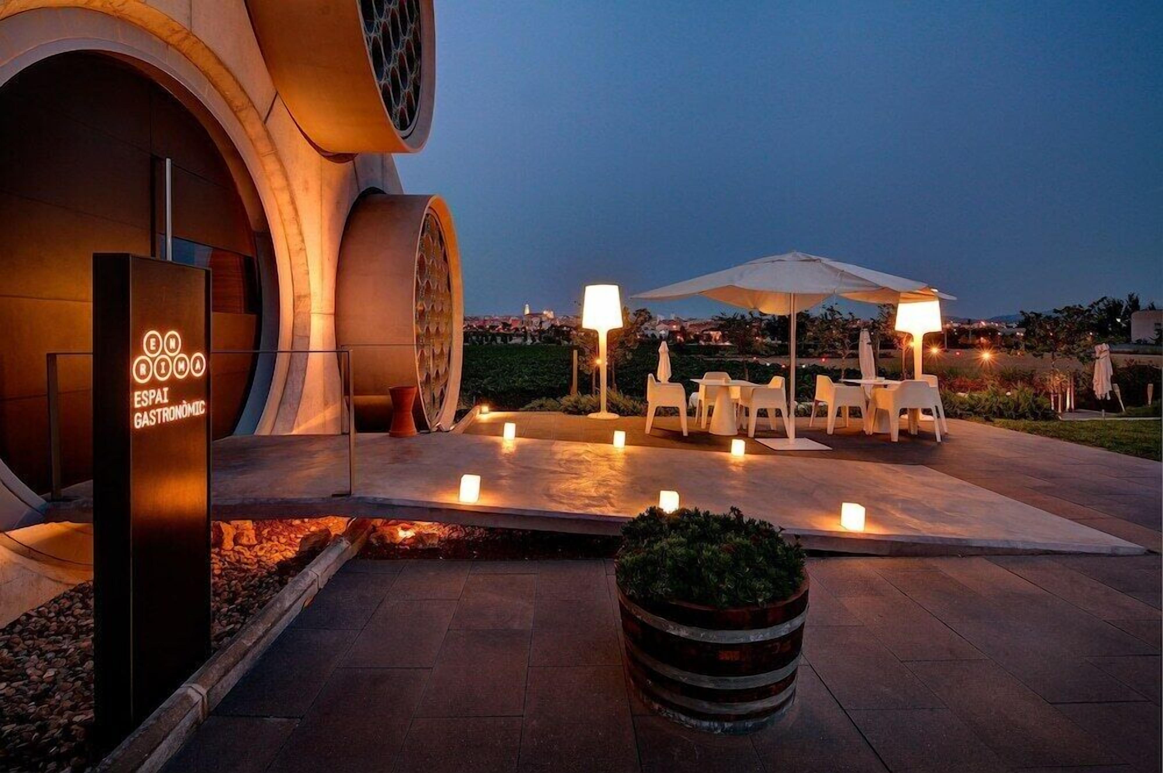 Un área de comedor al aire libre con sillas y mesas blancas, un paraguas grande y velas en el suelo.