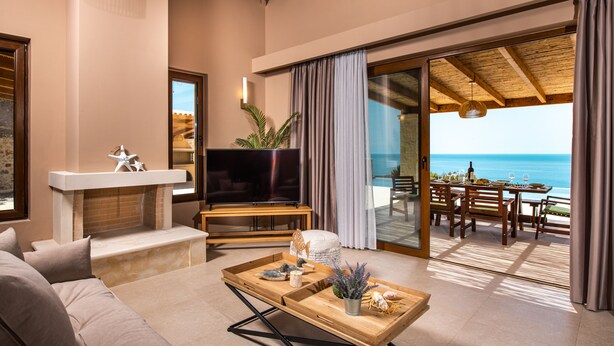 A modern living room with a sofa, a TV, a wooden coffee table, and a balcony with a view of the sea.