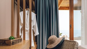 A room with a wooden vanity, a wicker chair, a white robe hanging on a wooden hanger, and a view of the sea through a large window.