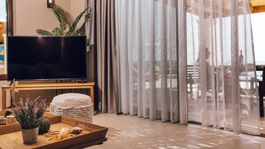 A modern living room with a flat-screen TV, a wooden coffee table with decorative items, and sheer curtains.