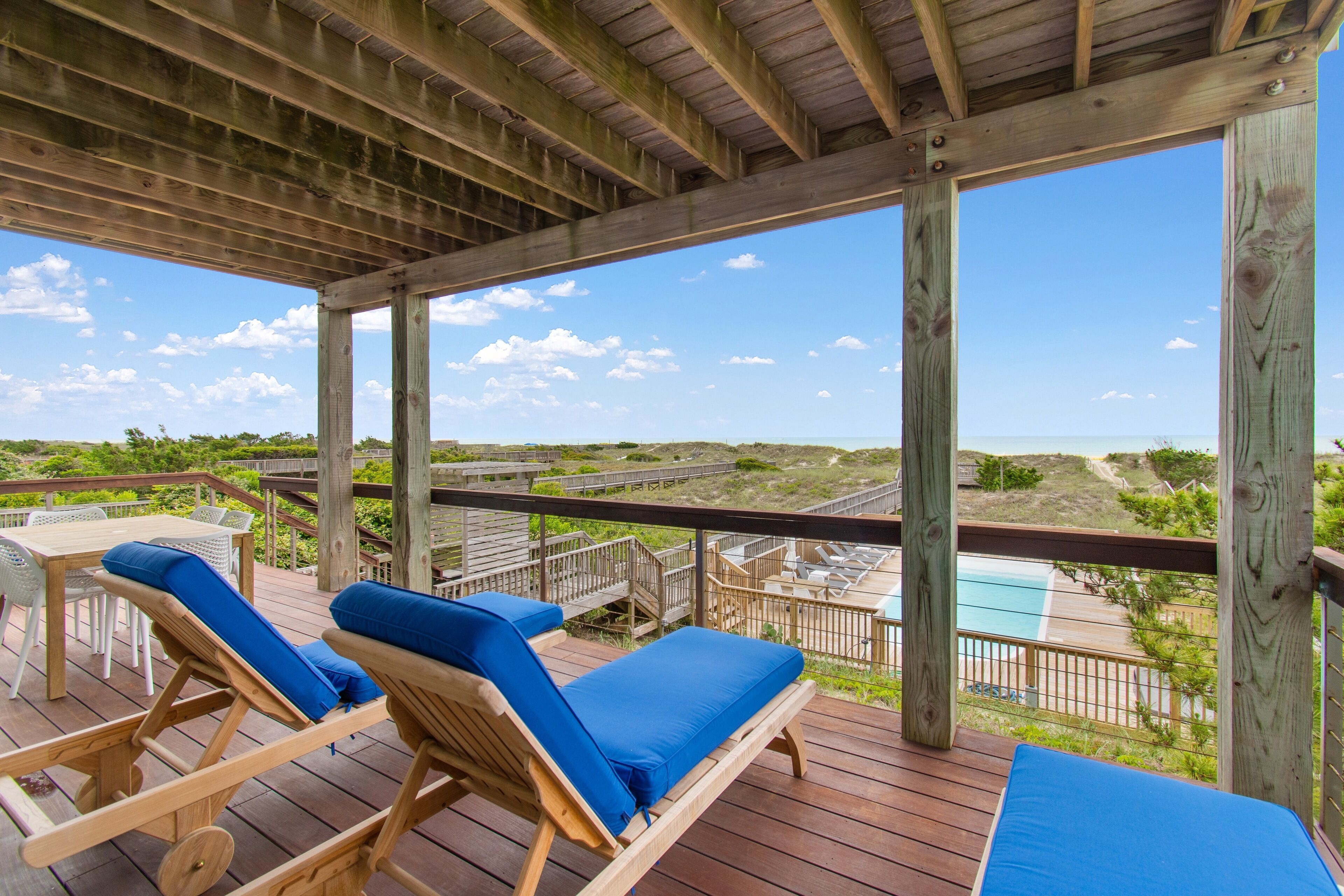 A wooden deck with two lounge chairs and a table, overlooking a beach and a pool.