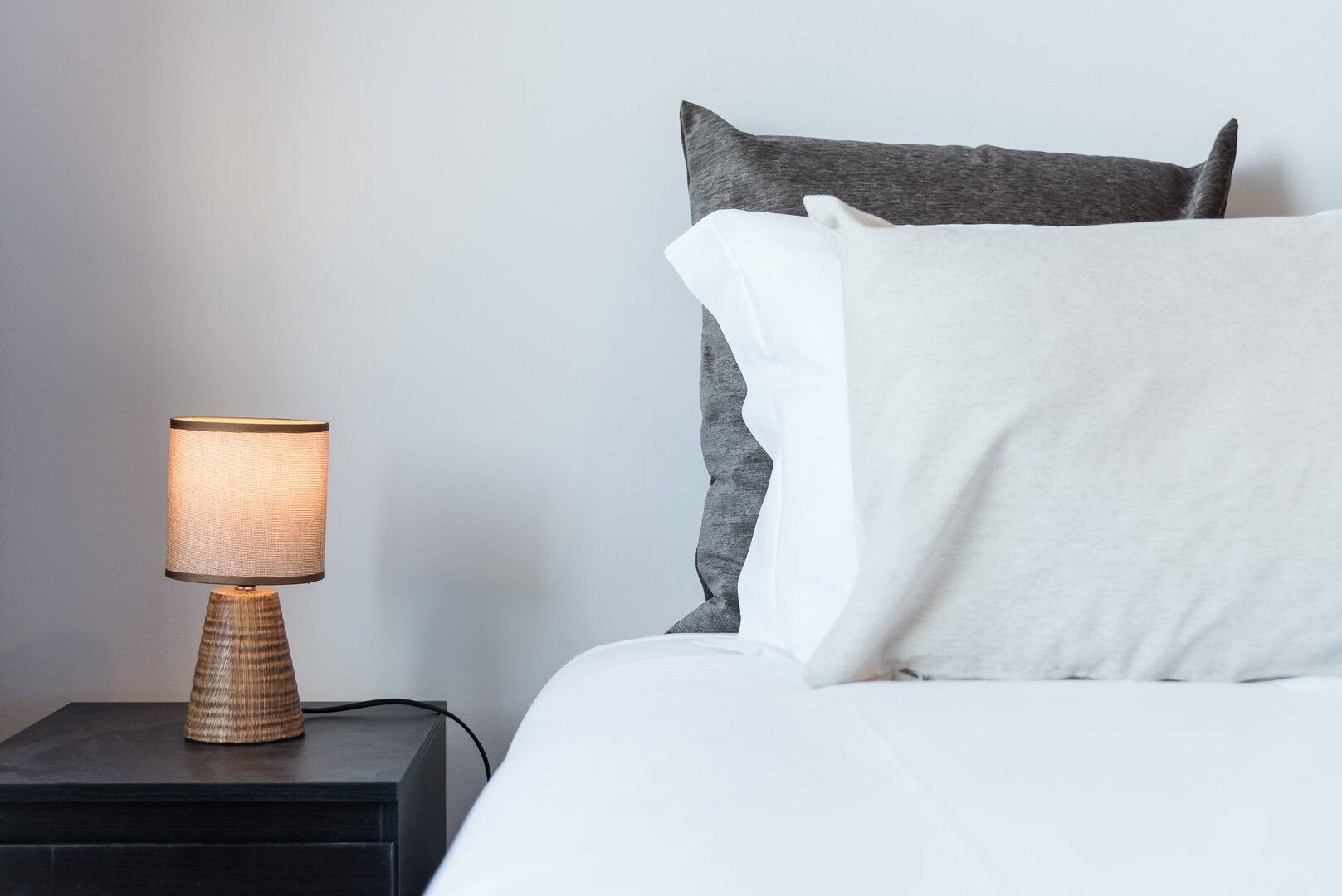 Ein Bett mit weißen und grauen Kissen, eine strukturierte Lampe auf einem schwarzen Nachttisch und eine schlichte Wand.