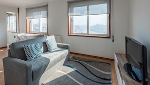 A modern living room with a grey sofa, two patterned cushions, a flat-screen TV, and large windows offering a city view. - GuestReady - Porto Magic View (Vila Nova de Gaia)
