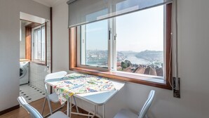 A small, bright room with a large window offering a city view, a table with a colorful tablecloth, and a washing machine in the corner. - GuestReady - Porto Magic View (Vila Nova de Gaia)