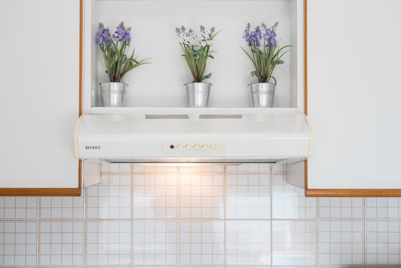 A kitchen with a white range hood, three potted plants, and white tiled backsplash.
