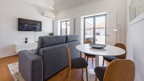 Un salon moderne avec un canapé gris, une table de salle à manger ronde entourée de chaises, un téléviseur à écran plat fixé au mur et une grande fenêtre offrant une vue sur l’extérieur.