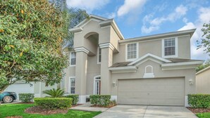 A two-story house with a beige exterior, a garage, and a well-maintained front yard with a lawn and shrubs.