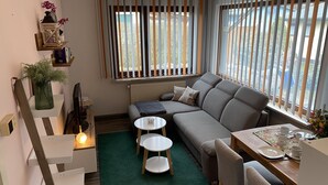 A modern living room with a sofa, two small tables, a green rug, and a window with vertical blinds.