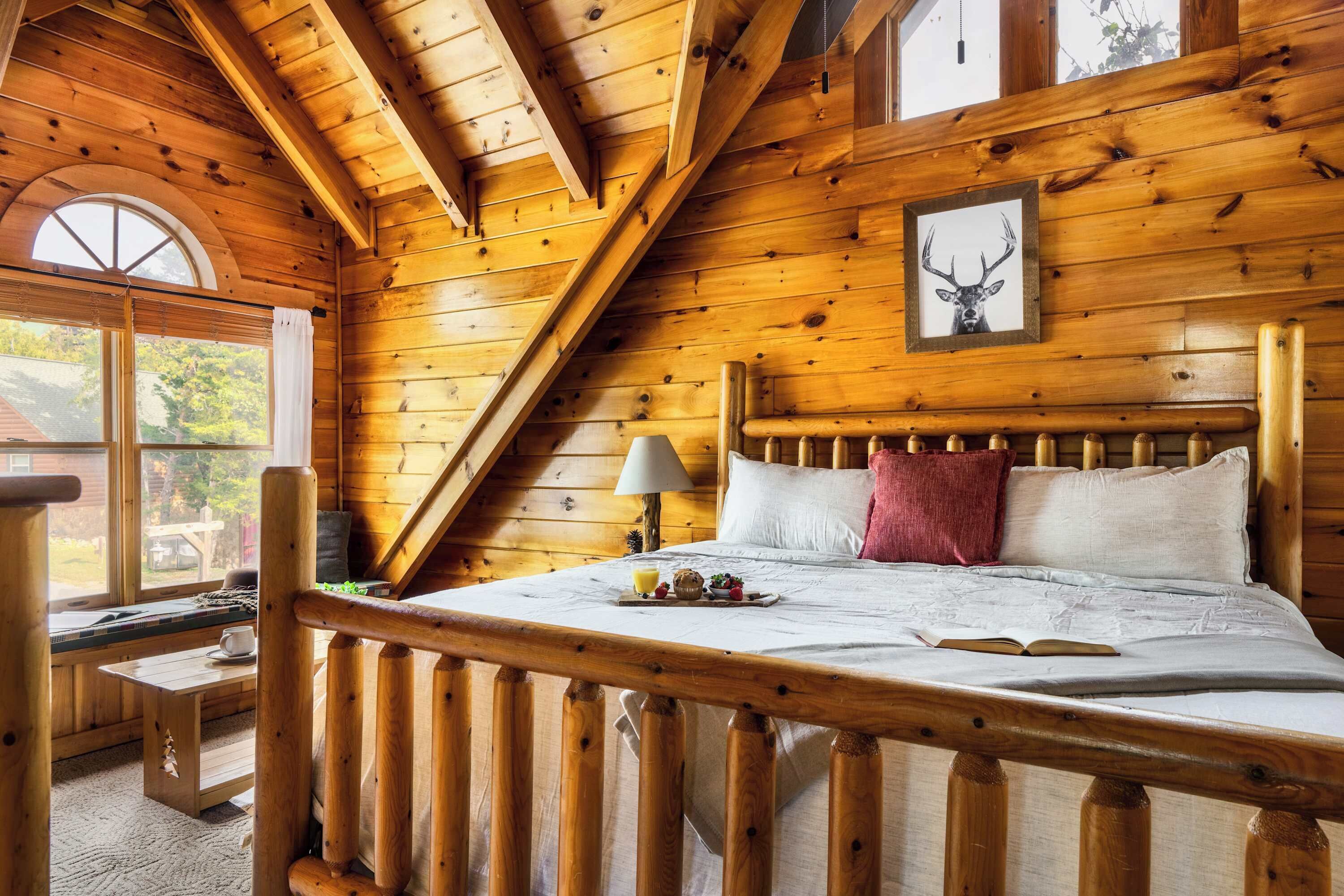 A cozy wooden cabin bedroom with a bed, pillows, a lamp, and a framed deer picture on the wall.