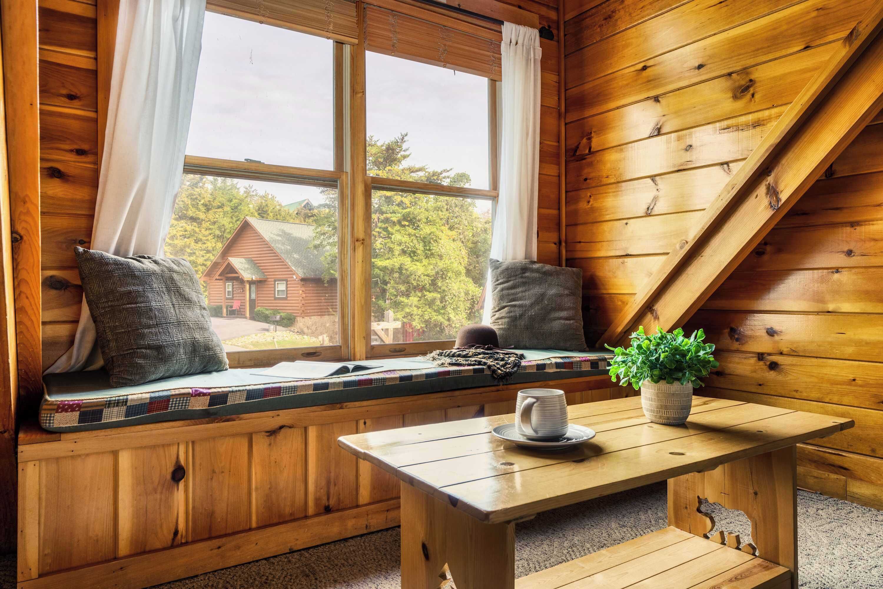 A cozy cabin interior with a wooden bench seat by a large window, a small table with a cup and saucer, and a potted plant.