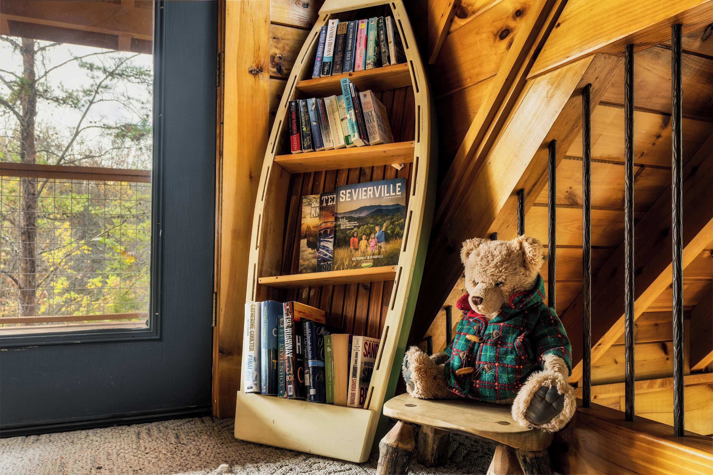 A wooden boat-shaped bookshelf filled with books, a teddy bear in a plaid jacket, and a window with a view of trees.