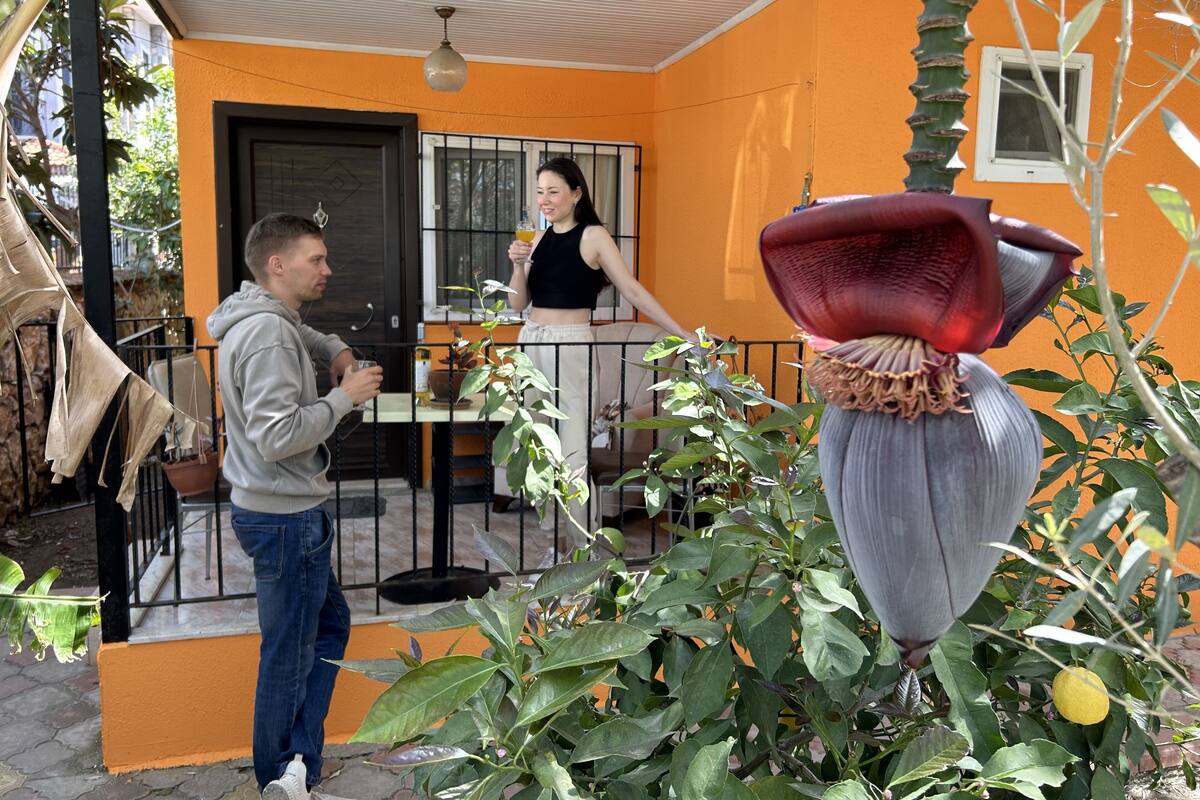 Un homme et une femme se tiennent devant un bâtiment à la façade orange, avec une porte noire et une fenêtre. Au premier plan, on aperçoit un pot de fleurs contenant une plante à feuilles vertes et une grande fleur rouge.