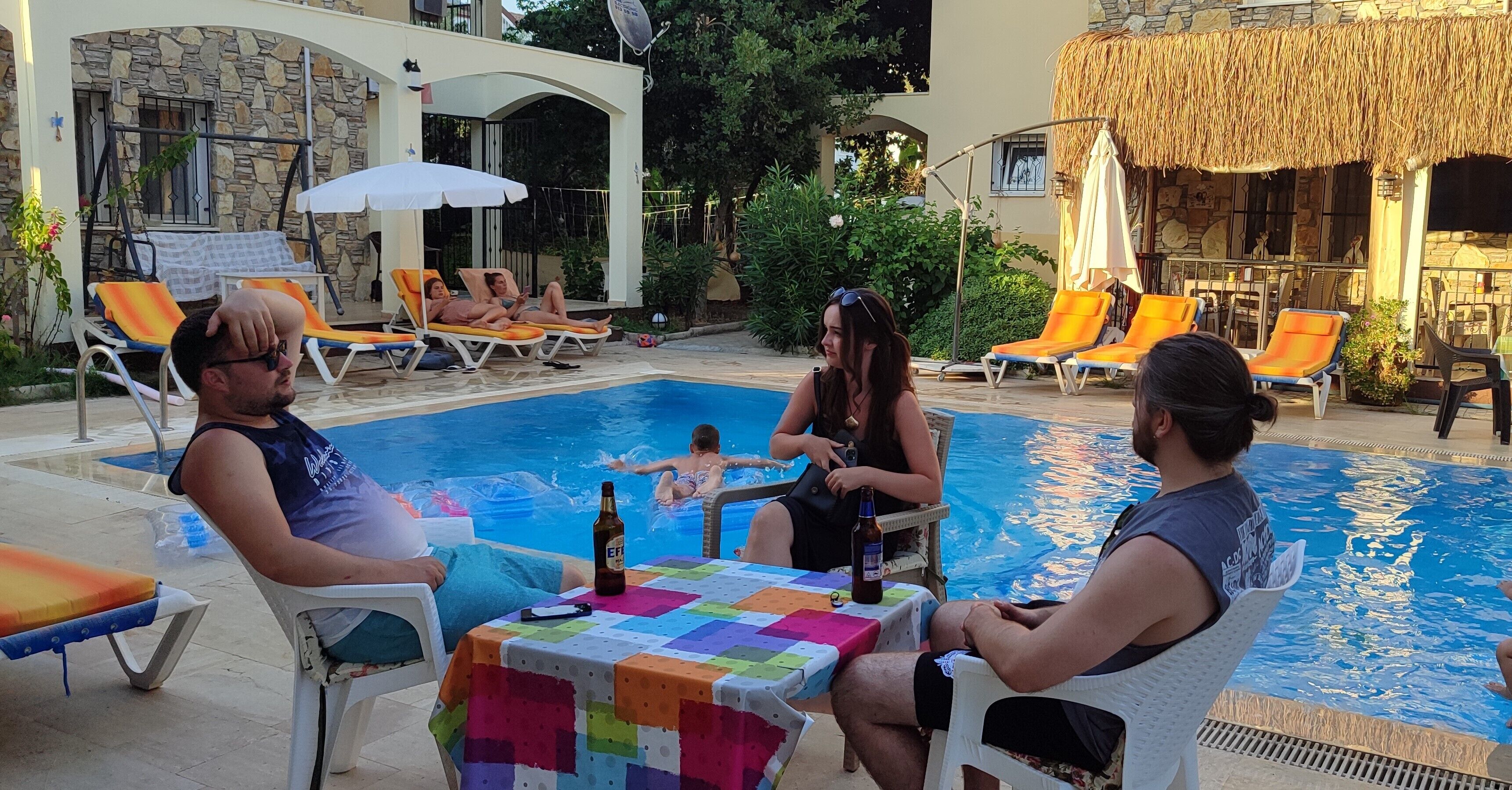 Un espace aménagé au bord de la piscine, avec des chaises longues, une table recouverte d’une nappe colorée et une bouteille de bière.