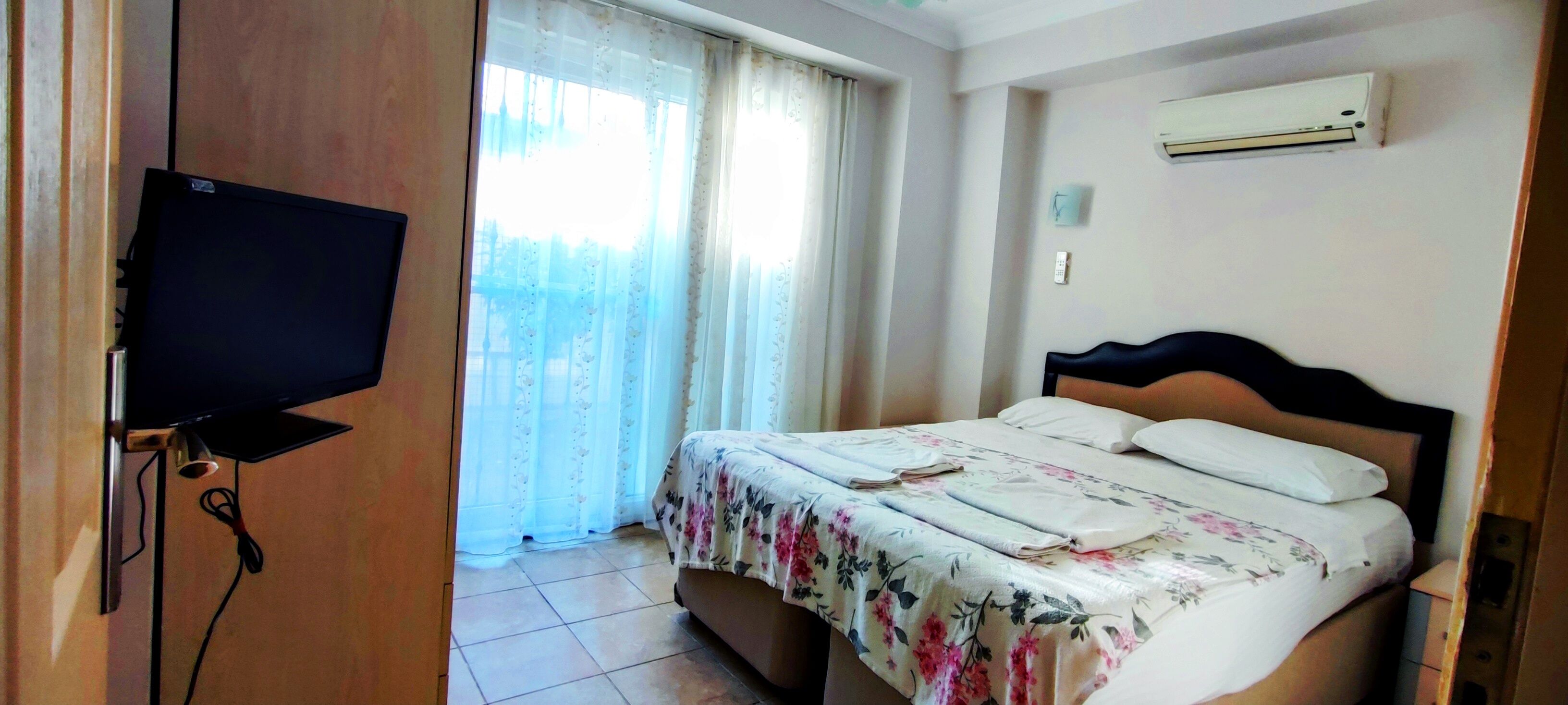Una habitación de hotel con una cama, televisión, ventana con cortinas y un equipo de aire acondicionado.