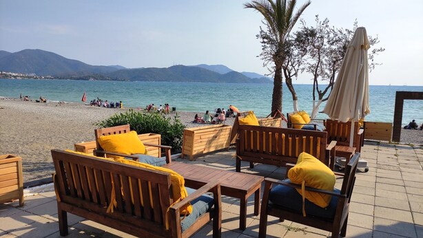 A beachside seating area with wooden furniture and umbrellas, overlooking a beach with people and mountains in the background. - Cileks Apart Otel (Fethiye)