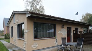 Une maison de plain-pied dotée d’un patio comprenant une table et des chaises, une petite construction en briques et une grande fenêtre.